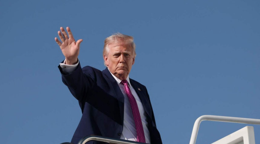 Donald Trump, dressed in a navy blue suit and purple tie, raises his right hand to wave from the steps of Air Force One against a clear blue sky. This strong gesture of leadership signifies the mood behind "Trump’s Unilateral Vow Opening the Strait of Hormuz," as discussed in this report.